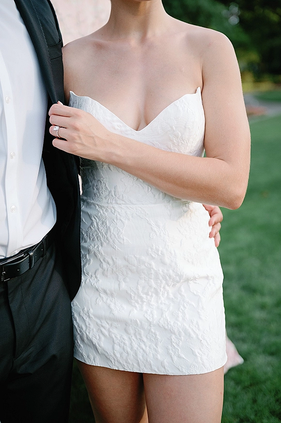 Wedding dress close-up of a strapless wedding dress with floral jacquard fabric, bride holding groom’s arm showing ring set, on green lawn with trees
