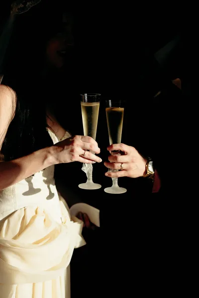 Wedding toast as bride and groom clink champagne flutes, rings and wristwatch visible against a dark indoor reception backdrop
