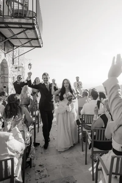 Wedding recessional as bride and groom walk the aisle, bride holding a bouquet, guests clapping on an outdoor terrace by a stone villa