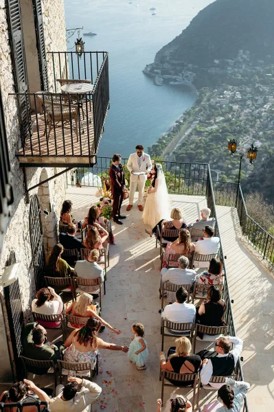 Wedding ceremony with bride and groom beneath a floral arch, long veil flowing as guests sit on a cliffside terrace above the ocean