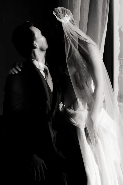 Wedding couple portrait in black and white, bride wearing veil and leaning over seated groom in suit, lit by window light and curtains