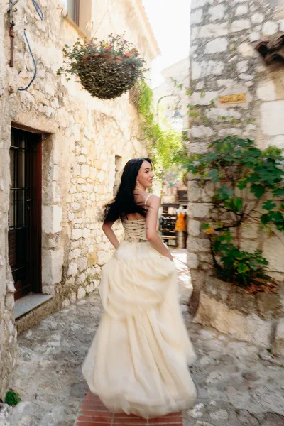 Bridal portrait of a bride walking away and looking back in a corset-back gown, long dark hair, in a cobblestone stone alleyway.