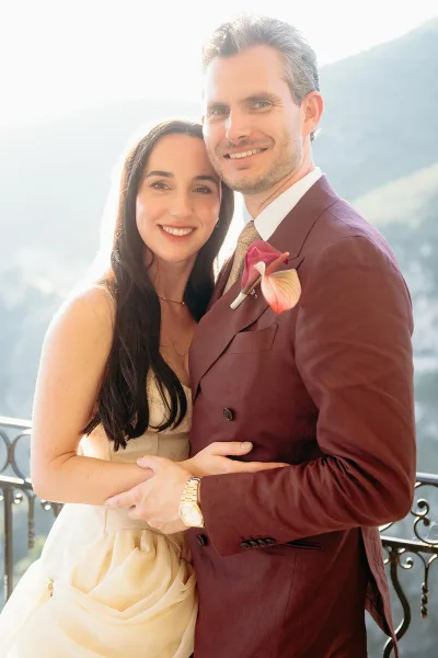 Couple portrait of bride and groom embrace on a balcony with a mountain lake view, bride in strapless gown, groom in burgundy suit