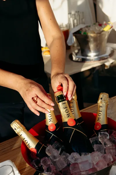 Champagne on ice in a metal bucket, champagne bottles nestled in ice cubes as a manicured hand reaches from a bar counter station