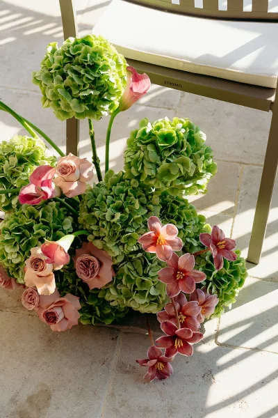 Ceremony aisle flowers with green hydrangeas and blush roses beside an aisle chair, accented by orchids on a sunlit stone tile floor