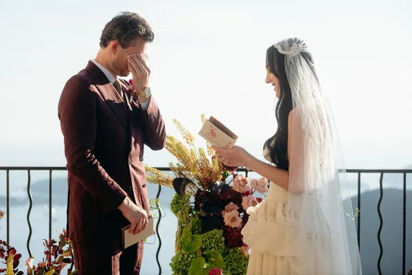 Wedding vows as bride reading vows from a vow book to her groom in a burgundy suit, long veil flowing on an ocean-view balcony