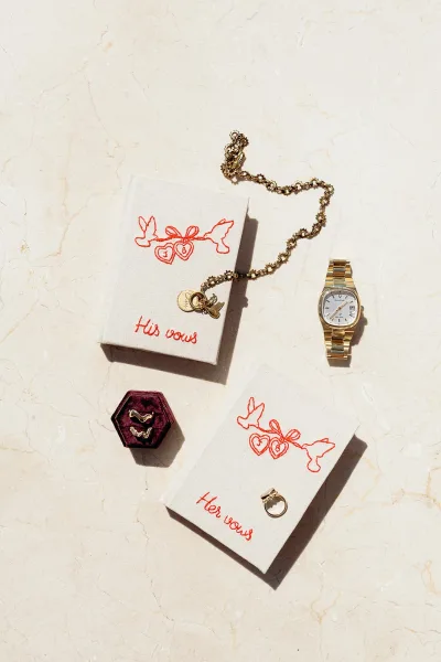 Wedding vow books in a his and hers vow books flat lay with embroidered covers, rings, gold watch, and velvet ring box on marble surface
