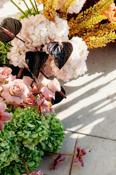 Wedding florals featuring hydrangea wedding flowers with blush roses and orchids arranged on a stone floor in sunlight shadows, gold foliage accents