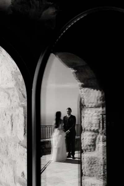 Couple portrait in a black and white wedding photo, bride and groom holding hands under a stone archway on a terrace balcony