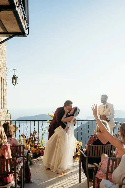 Ceremony kiss moment as bride in strapless gown and long veil kisses groom in burgundy suit, officiant behind on ocean-view terrace