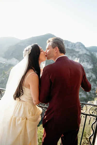 Wedding kiss as bride and groom kiss with lace-trim veil and champagne flutes on a balcony terrace with mountain view behind