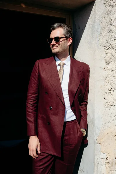 Groom portrait in a burgundy groom suit with sunglasses and tie, standing in a shadowed doorway against a rustic stone wall backdrop