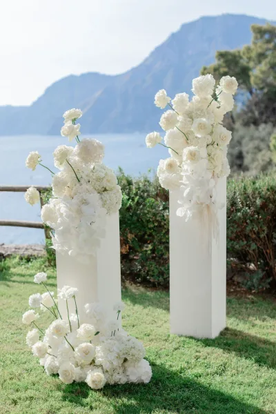 Wedding ceremony flowers on pedestal columns with white hydrangeas and roses, framing a lakeside lawn with mountains in the distance