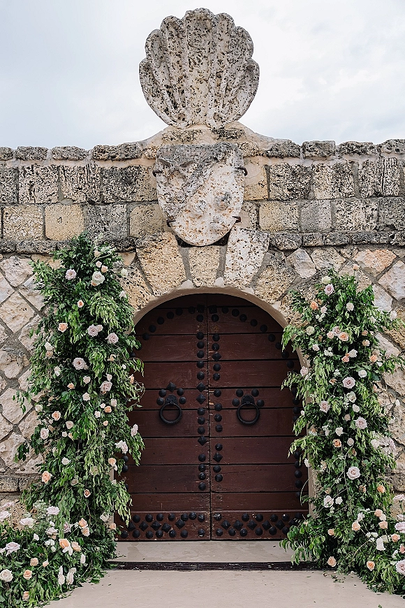 Wedding entrance decor with floral doorway arch of blush roses and greenery garlands framing arched wooden doors on a stone wall