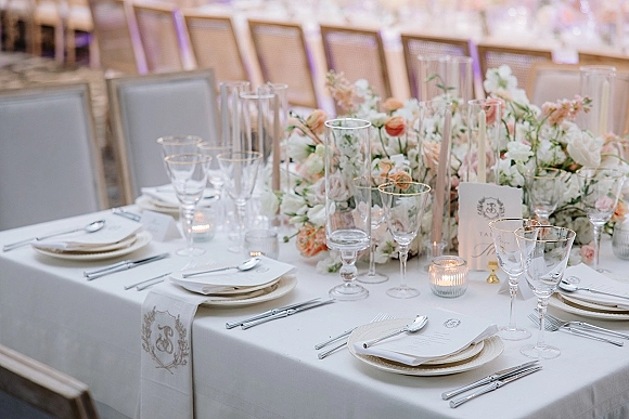 Reception tablescape with a wedding table setting of white linens, blush rose centerpiece, taper and votive candles, and gold-rimmed glassware in a ballroom