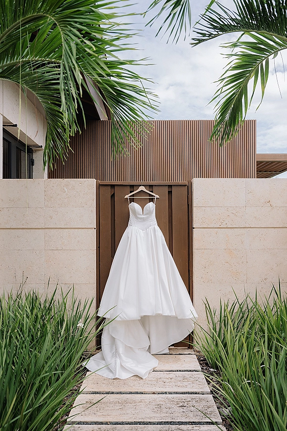 Wedding dress hanging on a wooden hanger, strapless satin gown with sweetheart neckline and long train against palm fronds and stone wall