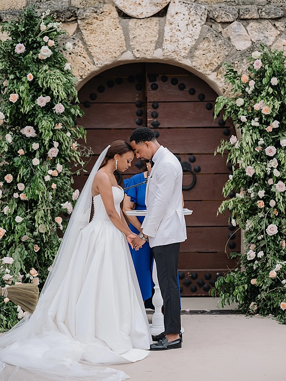 Wedding vows as bride and groom hold hands head to head under a rose and greenery arch by stone doors, veil flowing