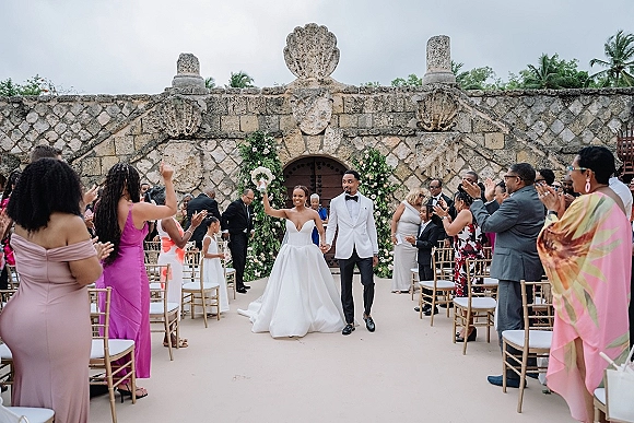 Wedding recessional as bride and groom walk the aisle hand in hand, bride lifting bouquet overhead in a courtyard with floral arch and stone wall backdrop