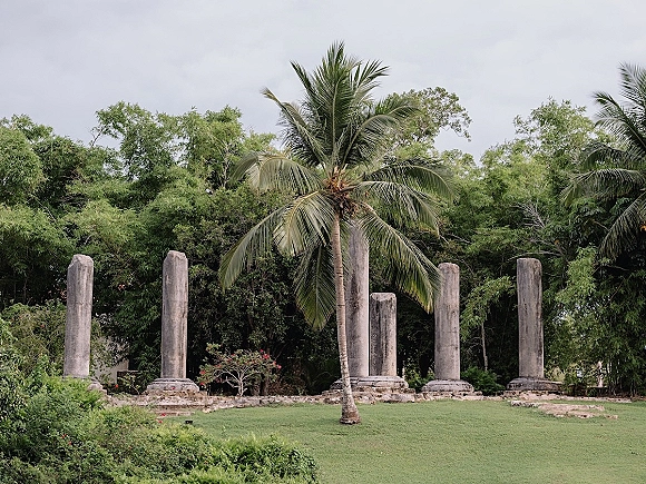 Outdoor photo backdrop with stone columns on a grassy tropical lawn, framed by palm trees and lush greenery under a cloudy sky