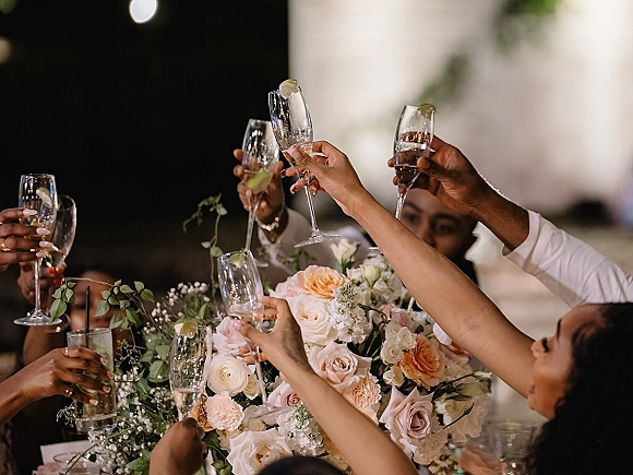 Wedding toast with raised champagne flutes and cocktail glasses around a rose centerpiece on a reception table under tent bokeh lights