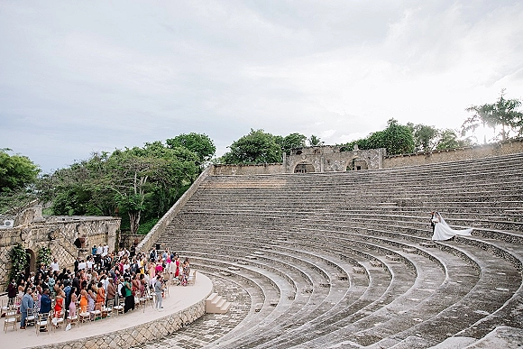 Wedding ceremony in a stone amphitheater with the bride in a flowing wedding dress and veil walking down steps toward guests in circle seating