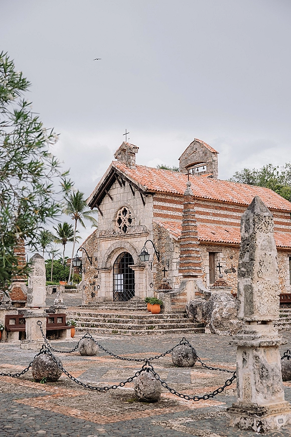 Stone chapel exterior with arched doorway and wrought iron gate, lanterns and terracotta roof tiles in a cobblestone courtyard with palms