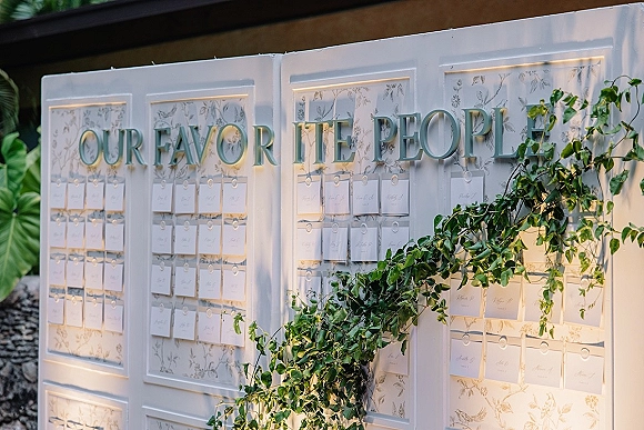 Wedding seating chart with escort card display on hooks, framed by greenery garland and large letter signage in sunlit outdoor setting