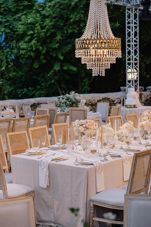 Reception tablescape with white linens, floral centerpieces, and taper candles under a crystal chandelier, set outdoors by trees and a stone wall