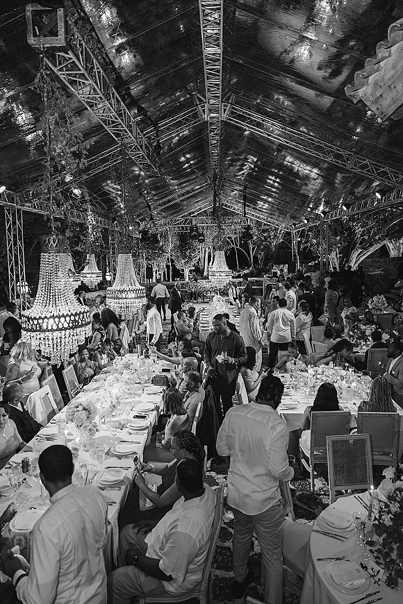 Reception tablescape with long banquet table wedding set in a clear-top tent, crystal chandeliers overhead, white florals and candles at night