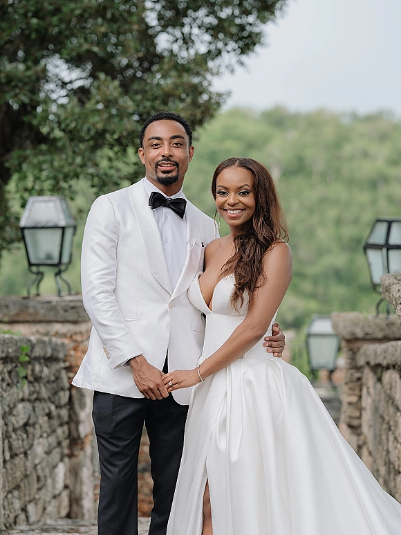 Couple portrait of bride in a strapless gown and groom in a white tuxedo holding hands and smiling by a lantern-lined stone wall outdoors