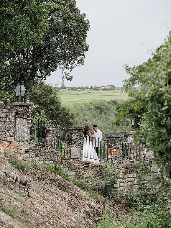 Couple portrait of bride and groom embracing on stone stairs, her long veil flowing by a wrought iron railing with lantern lights and greenery backdrop