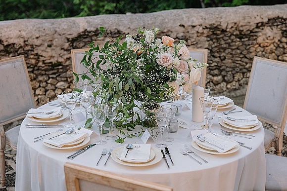 Reception tablescape with round wedding table setup featuring blush rose centerpiece, greenery garland, pillar candles, and stone wall backdrop