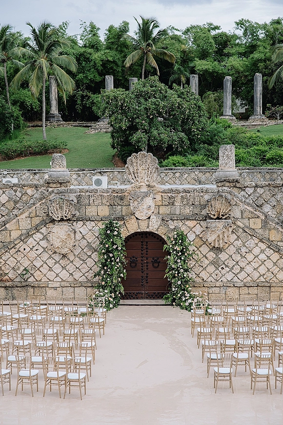 Ceremony setup with outdoor wedding ceremony chairs in gold chiavari rows and a blush floral arch by wooden doors on a stone terrace