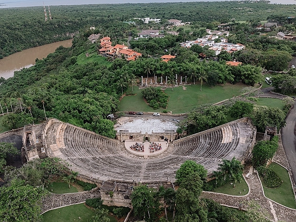Outdoor wedding ceremony in a stone amphitheater with guests seated around the stage on a green lawn beside a river and hills