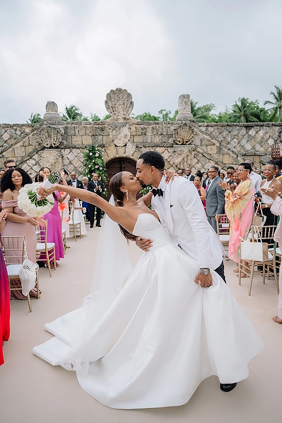 Wedding kiss portrait of the groom dipping the bride as she raises a white rose bouquet, cathedral veil flowing down the outdoor aisle by palm trees