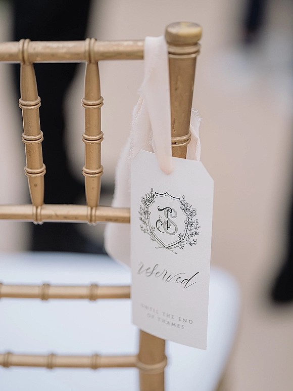Reserved seat tag with calligraphy and monogram crest, tied with ribbon to a gold Chiavari chair in a blurred reception space