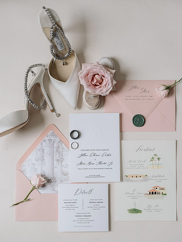 Invitation suite wedding invitation flat lay with blush envelopes, green wax seal, rings, and white crystal-strap heels on neutral fabric backdrop