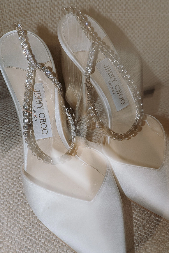 Bridal shoes in a wedding shoe flatlay, white satin pointed-toe slingback heels with crystal strap and chain detail on beige fabric