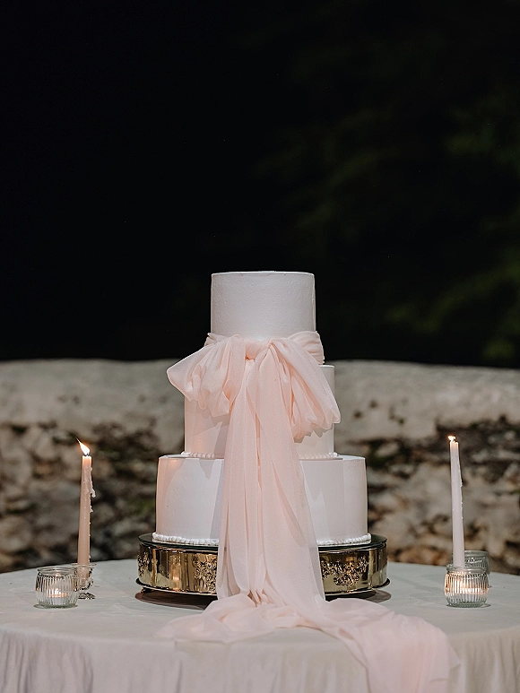 Wedding cake with smooth white buttercream tiers, styled on a stand with blush pink drape, taper candles, and stone wall backdrop