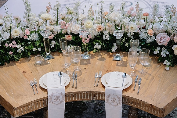 Reception tablescape with sweetheart table decor, lush blush and white florals, crystal candlesticks, and monogram napkins on wood table.