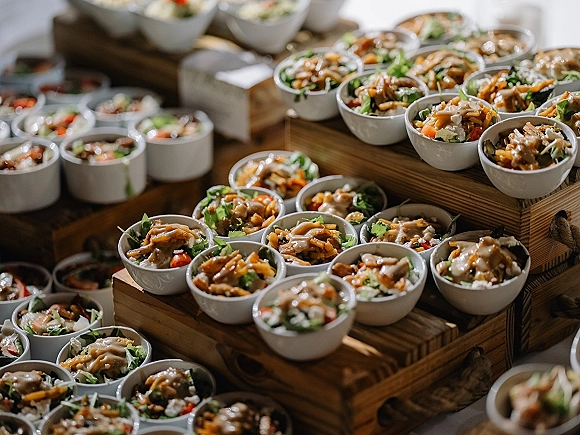 Wedding catering buffet display with mini salad bowls on rustic wooden crate risers, mixed greens and sliced vegetables on a tablecloth buffet table