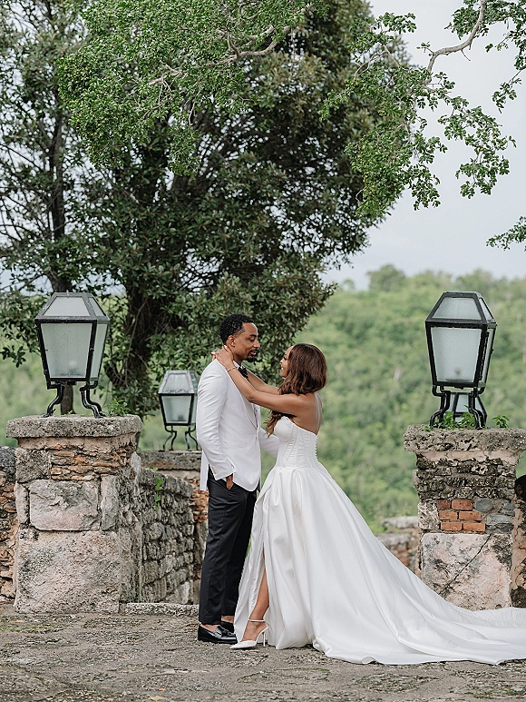 Couple portrait of bride and groom embrace on a stone terrace, her strapless gown with long train beside his white tuxedo jacket and lanterns