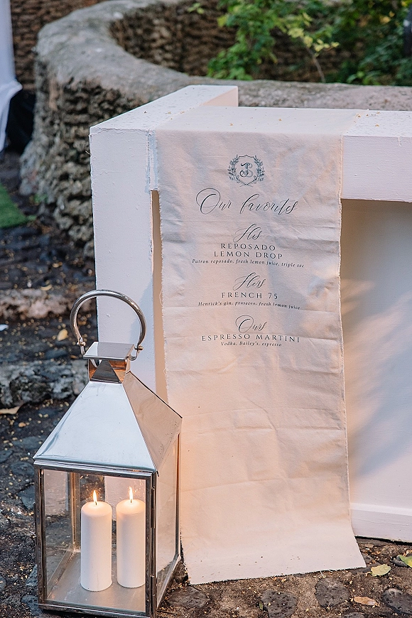 Wedding bar menu linen sign with calligraphy and crest monogram displayed beside a lantern and pillar candles on stone pavement outdoors