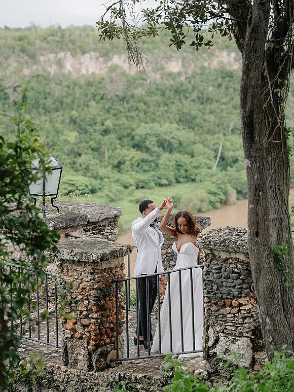 Couple portrait of bride in a strapless wedding dress twirling with groom in a white suit by iron railing on a stone terrace above a river view