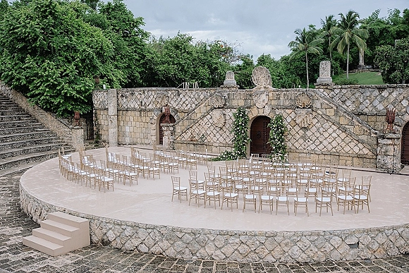 Ceremony setup with outdoor ceremony seating in a round layout of chiavari chairs around a floral arch in a stone courtyard with palm trees