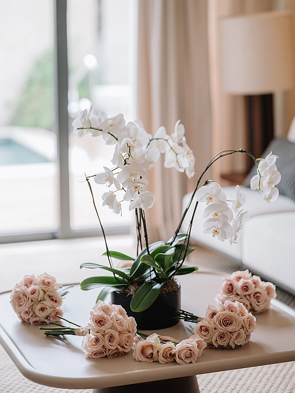 Bridal bouquets with blush rose bouquet clusters arranged on a white table beside a white orchid plant in soft window light indoors
