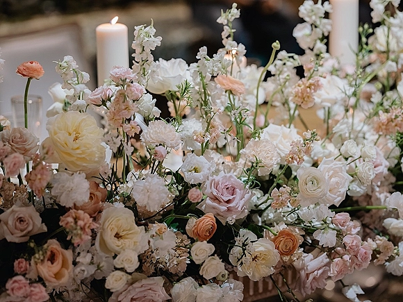 Wedding floral centerpiece with pastel roses and ranunculus, surrounded by taper candles and greenery on a dimly lit reception table
