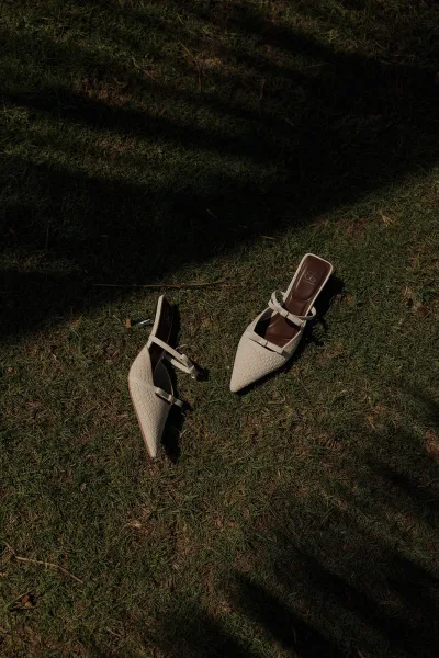 Bridal shoes, white bridal heels with pointed toes and ankle straps, arranged on grass lawn with soft tree shadows