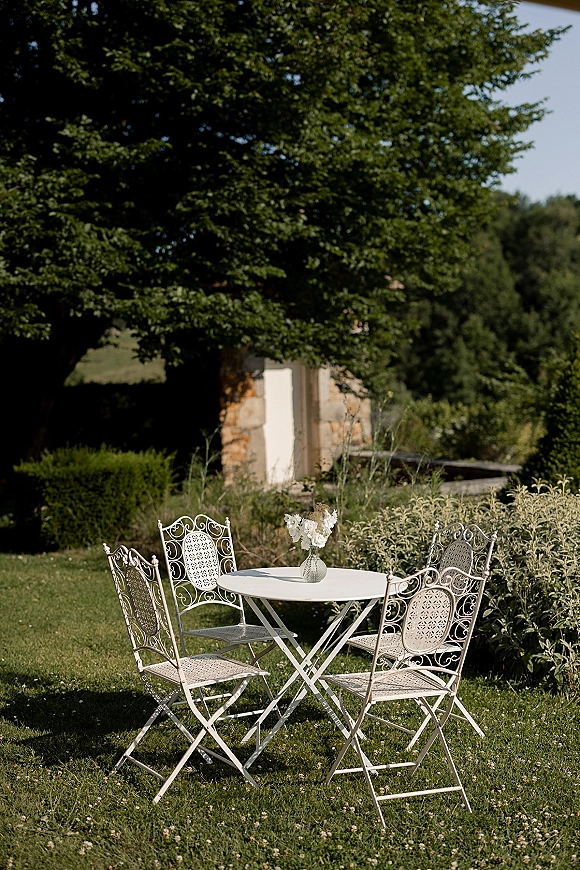 Garden seating vignette with wedding cocktail seating, a white metal bistro table and chairs with small vase of white flowers on a lawn by a stone building