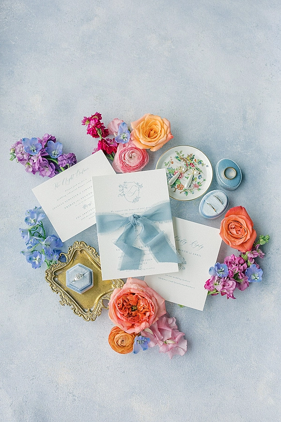 Invitation suite wedding invitation flat lay with blue ribbon, wax seal, floral sprigs, rings and pearl earrings on a light blue surface